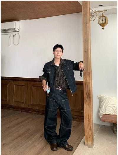 A man in a denim jacket and jeans stands in a room with wooden paneling and a wooden beam.