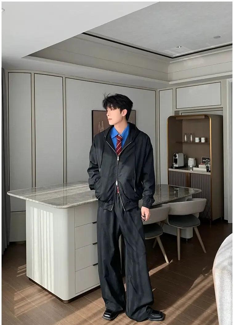 A man in a black suit stands in a modern, minimalist kitchen with a marble countertop and a glass dining table.