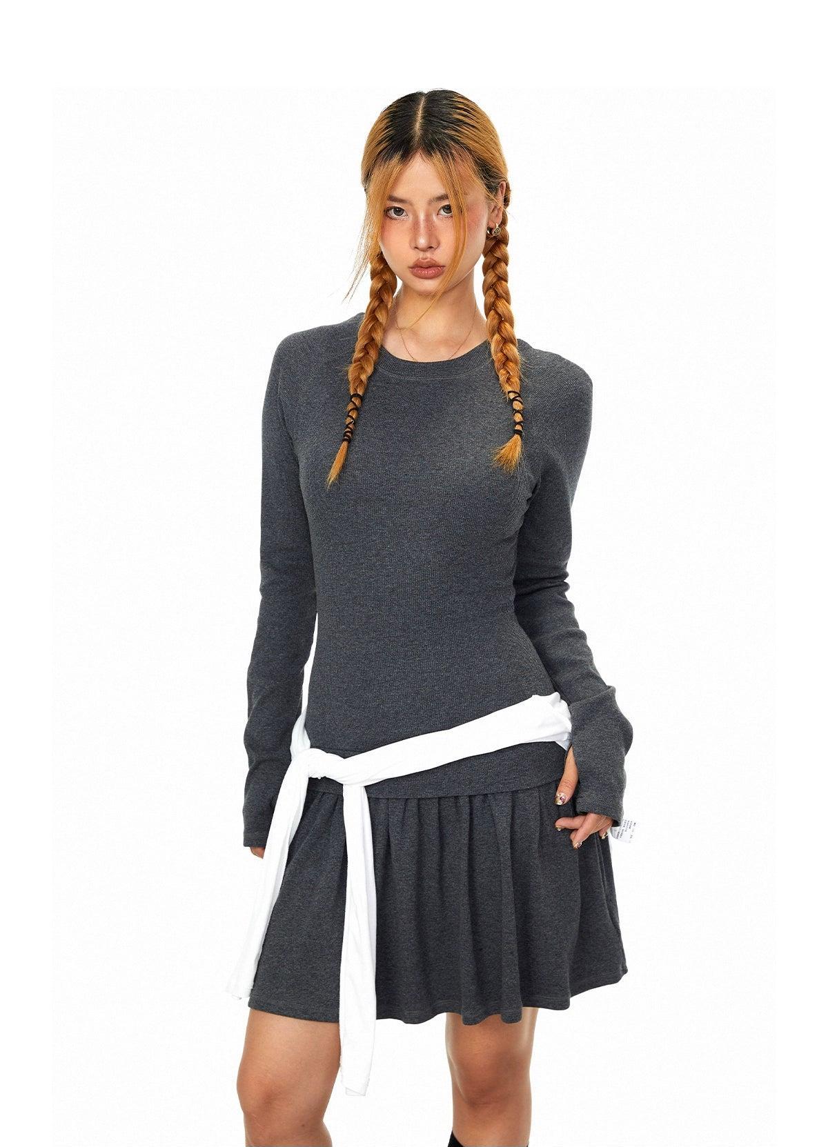 A young woman with long braided hair wearing a gray sweater and a gray skirt with a white belt, posing against a white background.