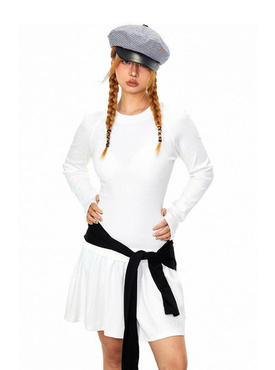 A woman wearing a white dress and a gray hat with a black band, standing with her hands on her hips.