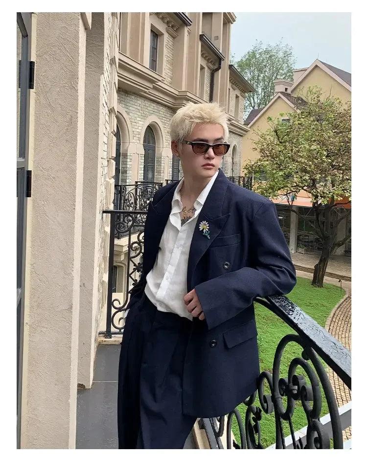 A person wearing a navy blue blazer and sunglasses is leaning against a wrought iron railing, with a building and trees in the background.