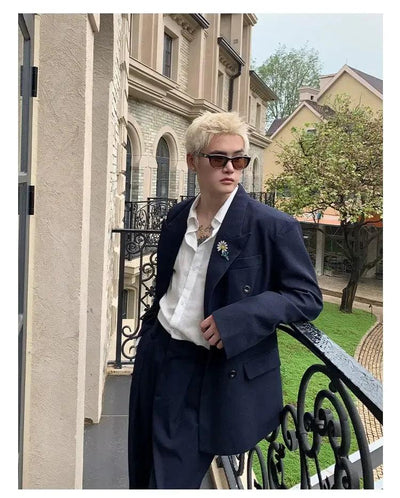 A person wearing a navy blue blazer and sunglasses is leaning against a wrought iron railing, with a building and trees in the background.