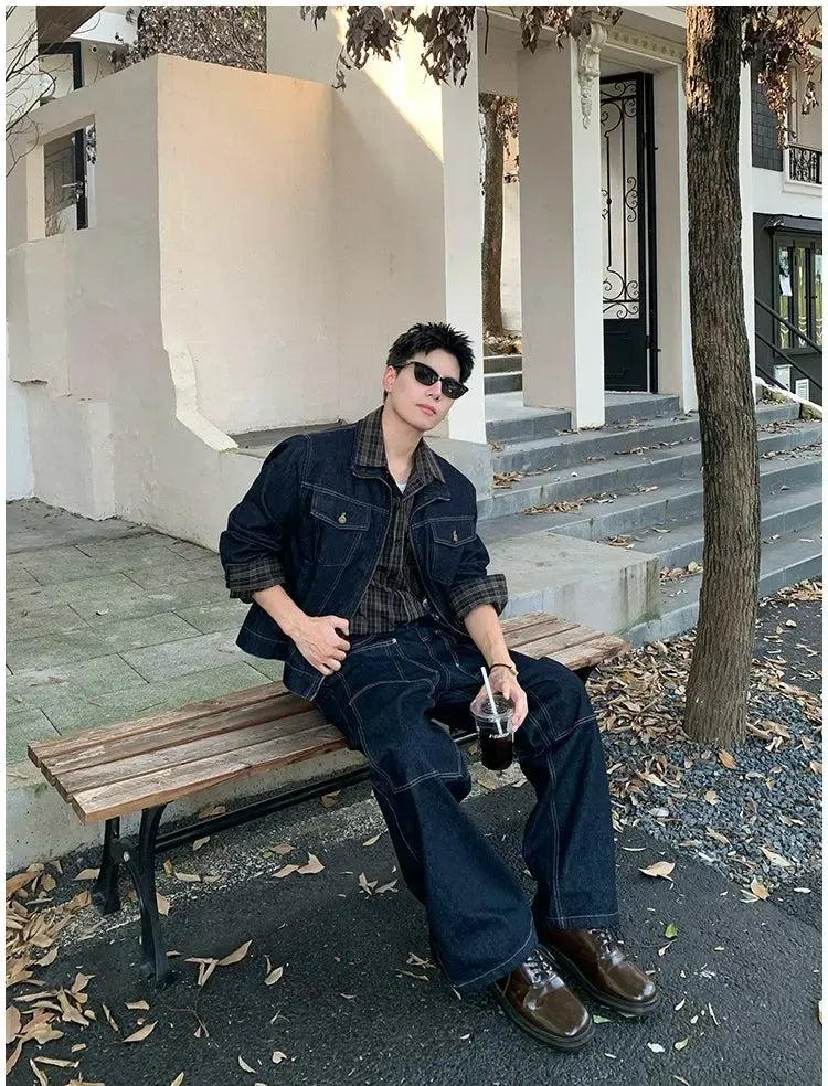 A person wearing a denim jacket and jeans is sitting on a bench, holding a drink, with a tree and a building in the background.