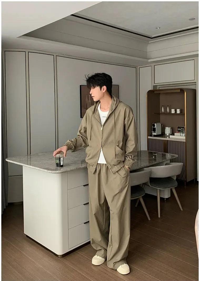 A person in a beige jacket and pants stands in a modern kitchen, holding a drink and looking off to the side.