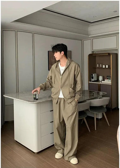 A person in a beige jacket and pants stands in a modern kitchen, holding a drink and looking off to the side.