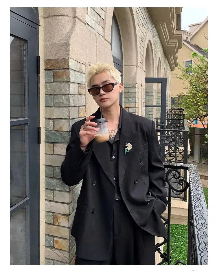 A person wearing a black suit and sunglasses is standing in front of a building, holding a drink in their hand.
