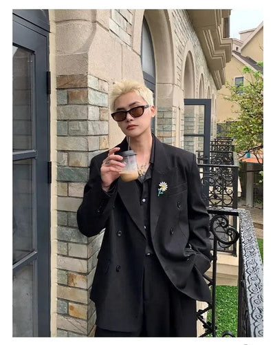 A person wearing a black suit and sunglasses is standing in front of a building, holding a drink in their hand.