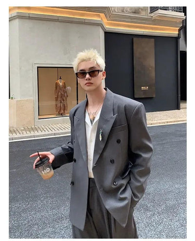 A young man wearing a gray suit and sunglasses is standing on a street, holding a drink in his hand.